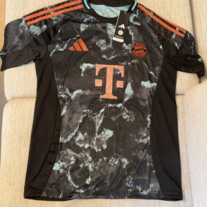 FC Bayern München Adidas fotbollströja - Snygg FC Bayern München fotbollströja från Adidas med svart bas och marmorerat mönster i ljusblått. Tröjan har korta ärmar, röda detaljer och klubbmärke på bröstet. Tillverkad i lätt och ventilerande material (AEROREADY) perfekt för träning eller match.