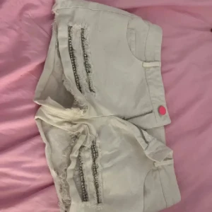 Vita lågmidjade shorts💗 - Snygga vita jeansshorts med råa kanter och slitningar framtill. Shortsen har två rader med glittrande strass på varje ben och en cool rosa knapp i midjan. Perfekta för dig som vill sticka ut lite extra i sommarvärmen!