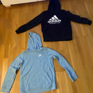 Svart Adidas och blå Nike hoodie - Två snygga hoodies: en svart Adidas Athletics med stor vit logga och en ljusblå Nike med liten vit logga på bröstet. Båda har huva och känguruficka, perfekta för chill dagar eller när du vill ha en sportig look. Båda har storleken 147 - 158 cm. 85kr styck. Båda för 150kr