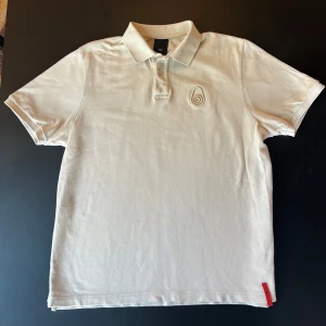 Beige Pike från Sail Racing - Snygg beige polo t-shirt från Sail Racing i bomull. Klassisk pikékrage med två knappar och broderad logga på bröstet. Röd detalj med varumärket nertill. Perfekt för en clean och sportig look.