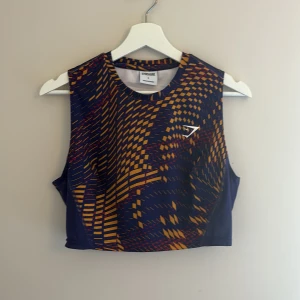 Mönstrad linnetopp från Gymshark - Säljer en cool linnetopp från Gymshark i storlek L. Toppen är marinblå med ett grafiskt mönster i gult och rött, har ärmlös design och croppad passform. Perfekt för träning eller när du vill sticka ut. Materialet är stretchigt och andas bra.
