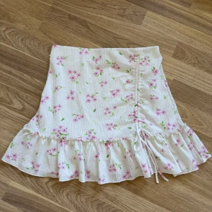 Blommig volangkjol från Gina Tricot - Supersöt kjol från Gina Tricot i vit med rosa blommor och gröna blad. Kjolen har volangkant nertill och en liten knytning på sidan. Perfekt för sommarens alla tillfällen och ger en fräsch och romantisk vibe. Aldrig använd.