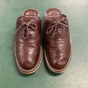 Bruna tofflor i broguemodell från Docksta - Stentuffa Dockstatofflor i äkta skinn med klassisk brogue-perforering och snörning. Modellen är öppen i hälen för en slip-on känsla. Perfekt för dig som gillar en mix av dressat och avslappnat. Nypris 1695:-