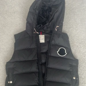 Svart dunväst från Moncler med huva - Säljer en svart dunväst från Moncler med stor vadderad huva och dragkedja framtill. Västen har ett stilrent Moncler-märke på bröstet och praktiska fickor med tryckknappar. Perfekt för lager-på-lager och riktigt snygg till streetwear.
