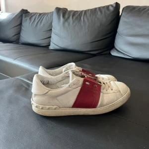 Valentino Garavani Open sneakers vit/röd - Snygga Valentino Garavani Open sneakers i vitt skinn med bred röd läderdetalj över mitten. Klassisk rund tå, platt sula och coola nitar baktill på hälen. Perfekta för dig som vill sticka ut med en lyxig touch och streetkänsla.
