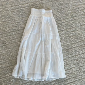 Vit långkjol från Gina Tricot - Superfin vit långkjol från Gina Tricot med smock i midjan och volangiga lager. Kjolen är luftig och har en flowy känsla, perfekt för varma dagar. Tillverkad i mjuk bomull som känns skön mot huden.