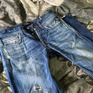 Blå slitna jeans från Replay, stl 27 - Säljer ett par blå jeans från Replay i storlek 27. Jeansen har slitna detaljer och hål på benen för en cool, avslappnad look. Klassisk femficksmodell med knappgylf och tydliga slitningar framtill. Perfekta för dig som gillar en edgy streetstil.