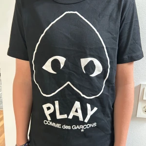 Comme des Garçons Play t-shirt - Svart t-shirt från Comme des Garçons Play med stort vitt tryck framtill, ikoniska hjärtögon och texten PLAY. Klassisk rund hals och korta ärmar. Skön passform och tillverkad i mjuk bomull. Perfekt för dig som gillar streetwear och stilrena prints. Kontakta mig vid minsta frågan