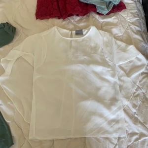  blus från Vero Moda i S - Superfin vit blus från Vero Moda i luftig och lätt polyester. Blusen har rund halsringning, korta vida ärmar och en snygg öppning med knapp i nacken. Perfekt när du vill ha en clean och stilren look.