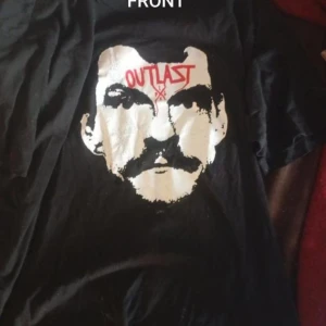 Christer Pettersson. Outlast merch. M - Svart t-shirt med stor vit och röd grafisk print på framsidan med texten 'OUTLAST' och ansikte. På ryggen finns en mindre logga med texten 'NORTHERN TYRANT' och grafiskt motiv. Klassisk passform och rund hals.