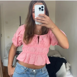 Rosa blus/topp med fina detaljer  - Supersöt rosa / korall färgad blus eller topp.  Kort ärm, volang nertill och smockad midja. Blusen har en liten knytning framtill och är croppad, perfekt till jeans eller kjol. Luftigt material som känns lätt och somrigt. Endast prövad, så i nyskick!