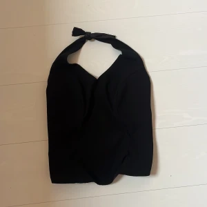 Svart halterneck korsett-topp - Snygg svart korsett-topp med halterneck och knyt i nacken. Toppen har en figurnära passform och smockad baksida för extra komfort. Perfekt för dig som vill ha en stilren och trendig look. Skulle uppskatta storleken som s/m 