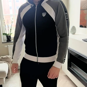 EA7 zip hoodie i svart, vitt och khaki - Snygg zip hoodie från EA7 Emporio Armani i svart, vitt och khaki. Jackan har huva med snörning, dragkedja framtill och coola EA7-loggor på bröstet, ärmen och ryggen. 