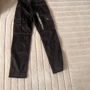 Svarta cargopants från Bluewear, stl 34 - Svarta cargopants från Bluewear med kontrasterande vita sömmar och flera praktiska fickor på sidorna. Byxorna har en rak passform och är tillverkade i slitstarkt bomullsmaterial. Perfekta för dig som gillar en cool och funktionell stil.