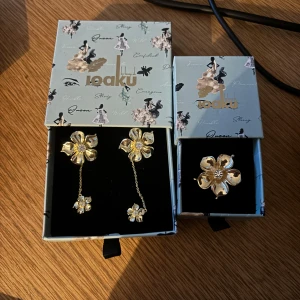 Guldiga blommiga örhängen & ring ioaku - Set med stora guldiga örhängen och matchande ring från ioaku. Både örhängen och ring har ett blommigt motiv med fem kronblad och en glittrig sten i mitten. Örhängena har en kedja med en mindre blomma som hänger ner för extra effekt. Allt är som i ny skick, örhängen: 450kr  Nypris 599kr, ring: 450 Nypris: 599kr. Pris vid köp av båda 800kr💕💕
