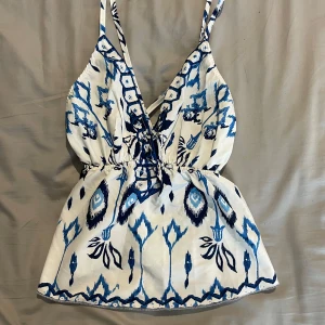 Vit och blå mönstrad linnetopp - Supersöt linnetopp med smala axelband och djup v-ringning. Toppen har ett blått och vitt mönster med inslag av olika blå nyanser och elastisk midja som ger en snygg passform. Perfekt för varma dagar och sommarvibbar!