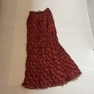 Mönstrad långkjol från Badal - Säljer en bohemisk långkjol från Badal i röd och orange med vågigt mönster och små cirklar. Kjolen har resår i midjan och är luftig med lätt volang nertill. Perfekt för dig som gillar färg och mönster! Skulle uppskatta storleken som storlek m