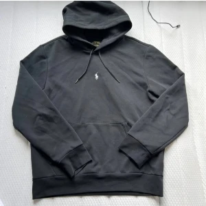 Svart Ralph Lauren hoodie - Svart hoodie från Polo Ralph Lauren med klassisk vit broderad logga på bröstet. Skön passform och tillverkad i mjuk bomullsmix, perfekt för chill dagar.