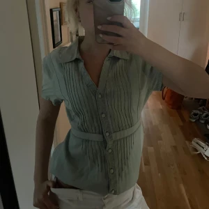 Jeansskjorta vintage - Superfin turkos ish jeansskjorta från gamla KappAhl med korta ärmar, knappar framtill och snygga veckade detaljer både fram och bak. Skjortan har en markerad midja med knytband i ryggen och klassisk krage. Säljs inte längre då det är en gammal design💕st.40 men passar mig som brukar ha s så skriver s