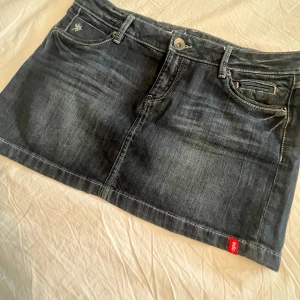 Mörk jeanskjol från edc by Esprit - Snygg mörkblå jeanskjol från edc by Esprit med klassisk femficksdesign, kontrastsömmar och broderade bakfickor. Kjolen har en kort modell och är perfekt för dig som gillar en tidlös denimlook. Liten röd logotagg framtill. Kjolen är lågmidjad.