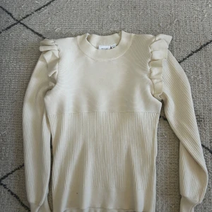 Beige stickad tröja med volang från Vila - Supersöt beige stickad tröja från Vila med volangdetaljer på axlarna. Tröjan har ribbad struktur både upptill och nedtill samt långa ärmar.