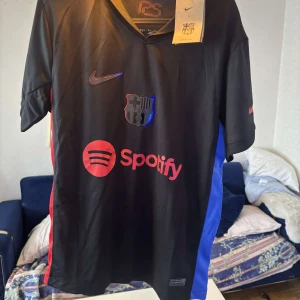 Svart FC Barcelona Nike fotbollströja - Snygg svart FC Barcelona fotbollströja från Nike med röda och blå detaljer på sidorna. Tröjan har klubbmärke och Nike-logga på bröstet samt Spotify som sponsor framtill. Tillverkad i lätt och ventilerande material, perfekt för match eller träning.