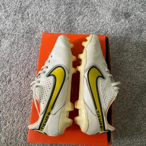 Nike Tiempo Legend 9 Pro FG fotbollsskor - Nike Tiempo Legend 9 Pro FG i vitt med gula och svarta detaljer. Skorna har snörning, rund tå och platt sula med fasta dobbar. Säljs på grund av att jag har vuxit ur och köpt nya, storlek 38.