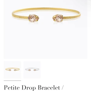 Guldfärgat armband Caroline Svedbom - Snyggt guldfärgat armband från Caroline Svedbom med två droppformade kristaller i ljus champagnefärg. Armbandet är öppet och har en elegant, tunn design med glittrig yta. Perfekt för att ge din look lite extra bling.