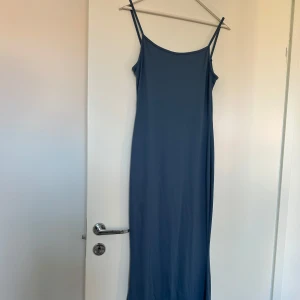Blå långklänning med smala axelband - En stilren blå långklänning med smala axelband och fin utsvängd passform. Klänningen är tillverkad i mjuk polyester och har en minimalistisk look som känns både modern och enkel att styla. Perfekt för dig som gillar en clean och avslappnad vibe.