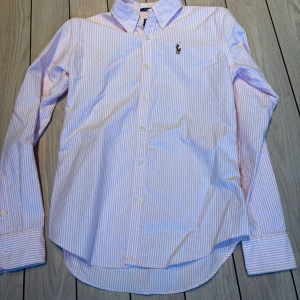 Rosa och vit randig skjorta Ralph Lauren - Snygg slim fit skjorta från Ralph Lauren i rosa och vitt randigt mönster. Klassisk button-down krage, långa ärmar och broderad logga på bröstet. Tillverkad i 100% bomull för en skön och fräsch känsla. Perfekt för dig som gillar stilrena och tidlösa plagg. Storlek 0, motsvarar ca XXS/32-34