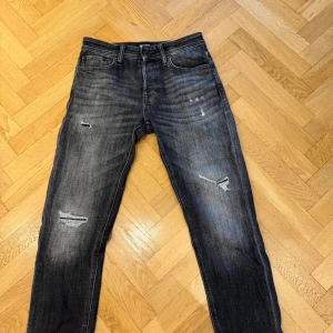 Svarta jeans med slitningar - Säljer ett par svarta jeans från Jack n Jones med schyssta slitningar på framsidan. Modellen Tapered/Mike.  Storlek W29 L32. Skick 9/10 använd fåtal gånger. Vid minsta fråga eller fundering hör av dig!