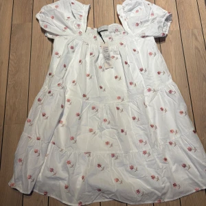 Vit blommig kortklänning Neo Noir - Supersöt vit kortklänning från Neo Noir med broderade rosa blommor, puffärmar och smockad överdel. Klänningen har en luftig passform och är gjord i bomull, perfekt för varma dagar. Romantisk och trendig look med volangdetaljer.