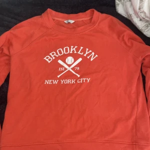 Röd/orange Brooklyn sweatshirt med tryck - Röd sweatshirt med vit text och basebolltryck på bröstet där det står 'Brooklyn New York City'. Tröjan har rund halsringning och långa ärmar. Perfekt för dig som gillar sportig och avslappnad stil.