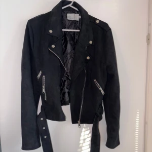 Svart mocka bikerjacka NLY Trend - Svart bikerjacka i mockaimitation från NLY Trend. Jackan har silvriga dragkedjor, knappar och bälte i midjan. Snygg krage och flera fickor med dragkedja ger en cool look. Perfekt för dig som vill ha en edgy stil.