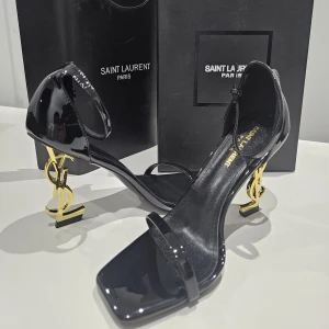 Svarta Saint Laurent klackar med YSL-häl - Exklusiva svarta klackar från Saint Laurent i lackat skinn med öppen tå och smal rem över foten. Snygg fyrkantig tå och ikonisk guldig YSL-logga som klack. Justerbar rem runt vristen för perfekt passform. Perfekta för dig som vill sticka ut med lyxig design.