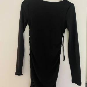 Svart tight klänning från Inawly - Svart kort klänning med lång ärm från Inawly. Klänningen är tight och har öppen rygg med snygg knyt-detalj upptill. Materialet är stretchigt och figurnära, perfekt för dig som gillar en stilren och enkel look med en twist.