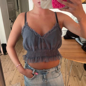 Grå volangig linnetopp med smock - Supersöt grå linnetopp med tunna axelband och volangdetaljer längs både urringning och nederkant. Toppen är croppad och har smockad midja som ger en snygg passform. Perfekt för varma dagar och att styla med jeans eller kjol.