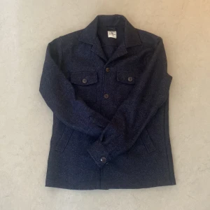 Mörkblå overshirt från John Henric - Snygg mörkblå overshirt från John Henric i ullblandning. Klassisk skjortkrage, två bröstfickor med knappar och raka sidfickor. Perfekt lager-på-lager-plagg med stilren look och rejäla knappar framtill. Passar dig som gillar en clean och tidlös stil.