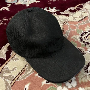 Svart Dior keps med monogram - Snygg svart keps från Dior med diskret monogrammönster över hela kepsen. Justerbar rem baktill i läder med Dior-logga. Klassisk böjd skärm och stilren look som lyfter vilken outfit som helst.