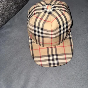 Burberry keps beige M - Snygg keps från Burberry i klassiskt beige rutigt mönster med svarta, vita och röda linjer. Märke på baksidan med texten 'BURBERRY LONDON ENGLAND'. Kepsen är tillverkad i bomull och polyester och har justerbar rem bak.
