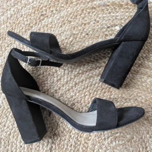 Svarta klackade sandaler från Nelly - Stilrena svarta sandaler från Nelly Shoes med bred klack och öppen tå. Skorna har en enkel rem över tårna och justerbart spänne runt ankeln. Tillverkade i mockaliknande material som ger en lyxig känsla. Perfekta för dig som gillar minimalistisk och trendig stil.