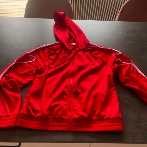 Röd vindjacka med hjärtdetalj från Monki - Röd vindjacka från Monki i storlek M, med huva och hel dragkedja framtill. Jackan har vita detaljer längs ärmarna och en unik hjärtformad kant på ena ärmen. Tillverkad i polyester och har ribbade muddar nertill och vid ärmslut.