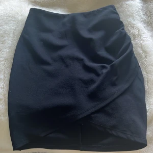 Svart omlottkjol från BikBok XS - Svart kjol från BikBok i omlottmodell med dragkedja bak. Går strax över knäna. Kjolen har en stilren look och är figurnära med lätt struktur i tyget. Perfekt för dig som gillar en enkel men snygg stil. 