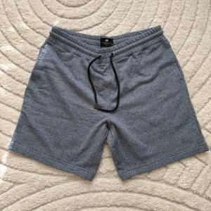 Grå mjuka shorts från H&M - Snygga grå shorts från H&M i mjuk bomullsmix med elastisk midja och svart snörning. Perfekta för chill dagar eller träning. Klassisk passform med fickor på sidorna och enkel stil som funkar till allt. Superbekväma och lätta att matcha med t-shirt eller hoodie.