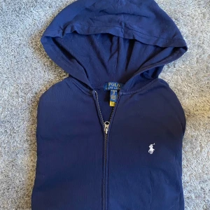 Marinblå hoodie från Polo Ralph Lauren - Snygg marinblå hoodie från Polo Ralph Lauren med dragkedja och klassisk vit broderad logga på bröstet. Tillverkad i 100% bomull för en skön känsla. Huva och ribbade muddar ger en avslappnad vibe. Perfekt för dig som gillar stilren streetwear. Storlek ca S/M 