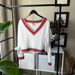 Vit ribbad stickad tröja Divided - Vit ribbad stickad tröja från Divided med v-ringad hals och sportiga ränder i rött och svart vid hals, ärmslut och nederkant. Croppad modell med lång ärm, perfekt för en chill och trendig look.