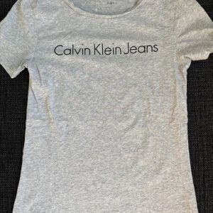 Grå Calvin Klein Jeans t-shirt - Snygg grå t-shirt från Calvin Klein Jeans med klassisk logga tryckt över bröstet. T-shirten har rund halsringning och korta ärmar. Tillverkad i mjuk bomull för en skön och avslappnad känsla. Perfekt till jeans eller shorts.