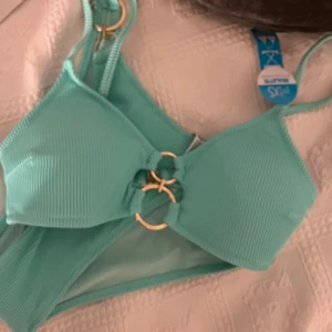 Turkos ribbad bikini med ringdetaljer (HELT NY, PRISLAPP) - Snygg turkos bikini från newyorker i ribbat material. Toppen har coola guldfärgade ringdetaljer framtill och smala axelband. Perfekt för strandhäng och poolparty! Fräsch och trendig färg som verkligen poppar.