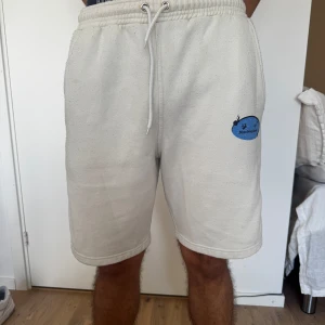 Beiga shorts med blått tryck och snörning - Snygga beiga shorts från Stretching Bends med elastisk midja och snörning. På ena benet finns ett blått tryck med logga och på baksidan en stor blå orm. Mjukt bomullsmaterial och avslappnad passform, perfekta för sommaren.