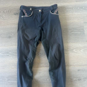 Mörkblå ridbyxor med detaljer - Snygga mörkblå ridbyxor från Cavallo med helskodd insida i mockaliknande material. Fickor med röd- och vitrandiga detaljer samt silvriga knappar. Broderad logga bak. Perfekta för dig som vill ha stil på ridbanan. Finns skavanker men inget tydligt😊står ingen storlek men skulle säga s